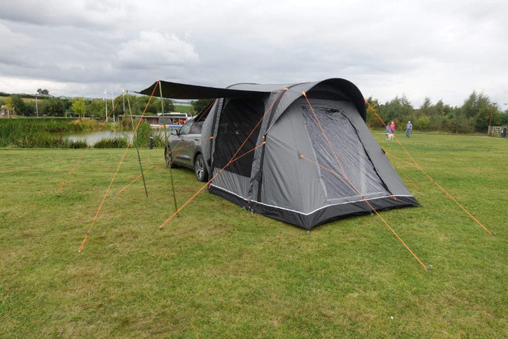 Camptech Adelaide rear-mounted inflatable car tent with grey fabric and orange ropes, pitched beside a lake under cloudy sky.