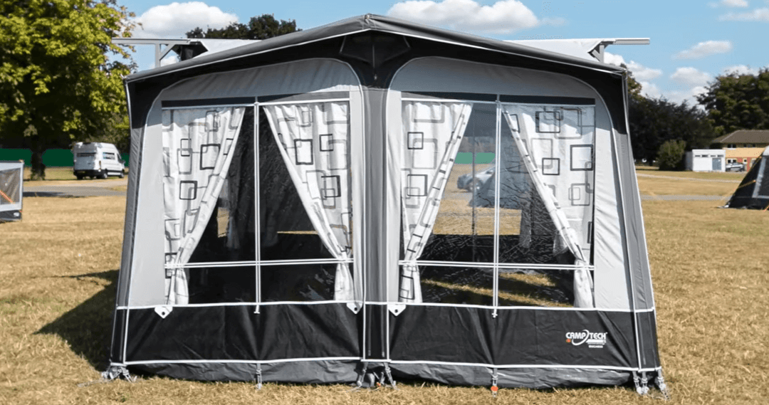 Camptech Duchess porch awning front view showing large clear panels and patterned curtains, set up on dry grass under sunny weather.