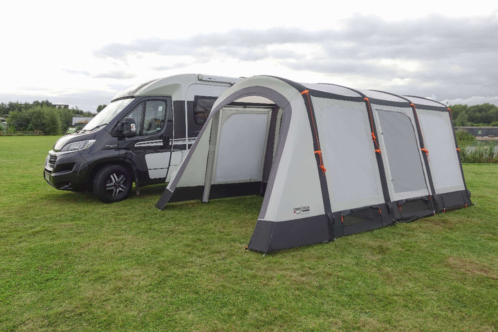 Camptech Moto Canterbury driveaway awning attached to a campervan on a green field, featuring grey and white panels with orange guy lines under a cloudy sky.