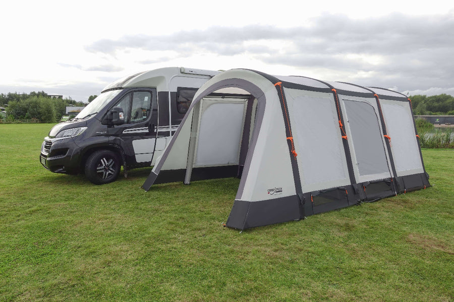 Camptech Moto Canterbury driveaway awning attached to a campervan on a green field, featuring grey and white panels with orange guy lines under a cloudy sky.