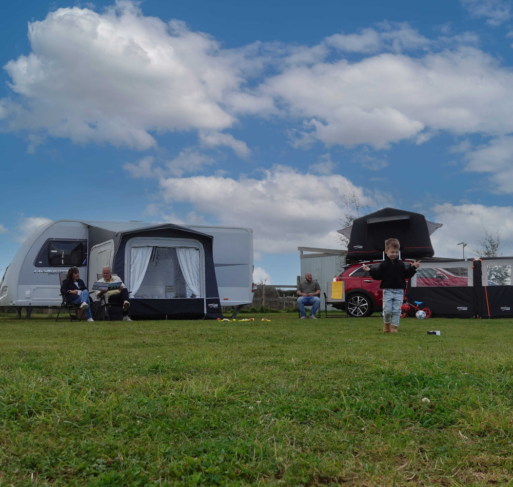 Family camping scene with Camptech Prince DL porch awning attached to a caravan, people relaxing outdoors with a car and awning setup in the background.