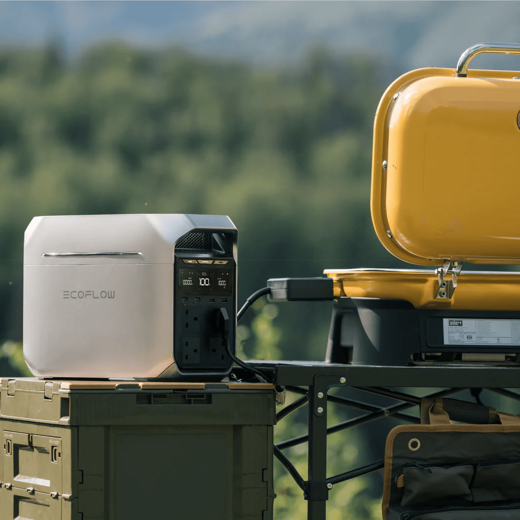 EcoFlow DELTA 3 Series placed outdoors on a green storage crate, plugged into a yellow electric grill during daytime, with a scenic forest and mountain landscape in the background.