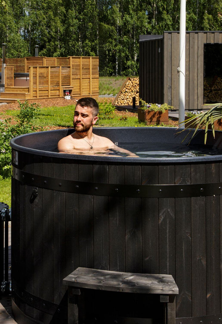 Person soaking in a Harvia wooden hot tub in a natural garden setting with modern cabin structures in the background.
