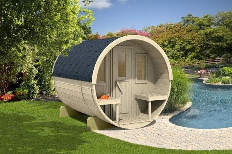 InMedias Barrel Sauna 250 in a landscaped backyard, positioned beside a pool with surrounding trees, showing its natural wood color and compact cylindrical design.