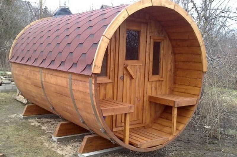Exterior view of InMedias Barrel Sauna 400 made of dark stained wood with a red shingle roof, standing on wooden supports in a garden  natural background.