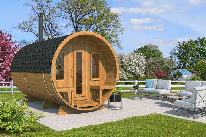 Outdoor view of InMedias Barrel Sauna 400 Deluxe placed on a patio, surrounded by modern garden furniture and springtime greenery, on a blue sky background.