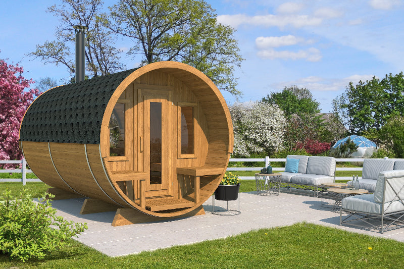 Outdoor view of InMedias Barrel Sauna 400 Deluxe placed on a patio, surrounded by modern garden furniture and springtime greenery, on a blue sky background.