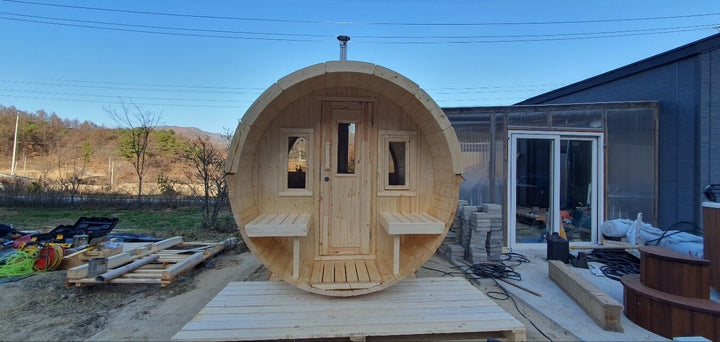 Front view of InMedias Barrel Sauna 250 under construction, showing a wooden cylindrical sauna structure with a central door and two side windows with small benches – background features blue sky and rural scenery.