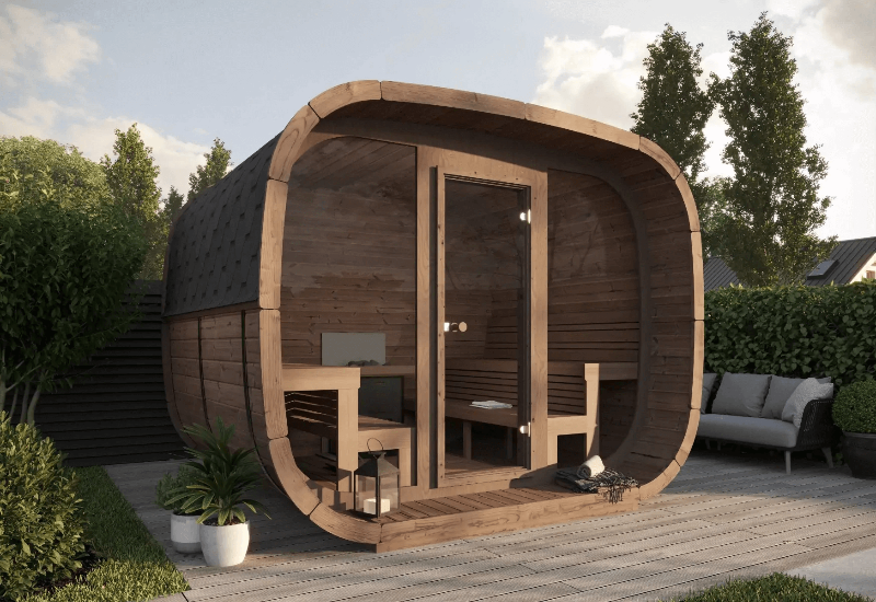 InMedias Cubus Premium Outdoor Sauna made of dark wood with a curved front and glass door, placed on a wooden deck with surrounding greenery and potted plants , on a  outdoor background.
