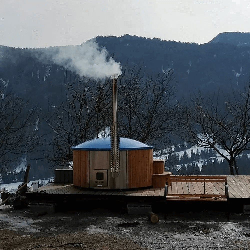 InMedias Delux 220 XL Hot Tub with a blue cover and chimney releasing steam, placed on a deck in a snowy mountain landscape with bare trees and distant hills.