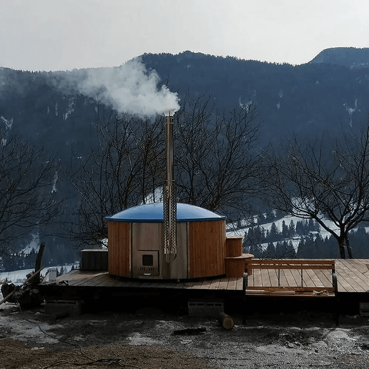 InMedias Delux 220 XL Hot Tub with a blue cover and chimney releasing steam, placed on a deck in a snowy mountain landscape with bare trees and distant hills.