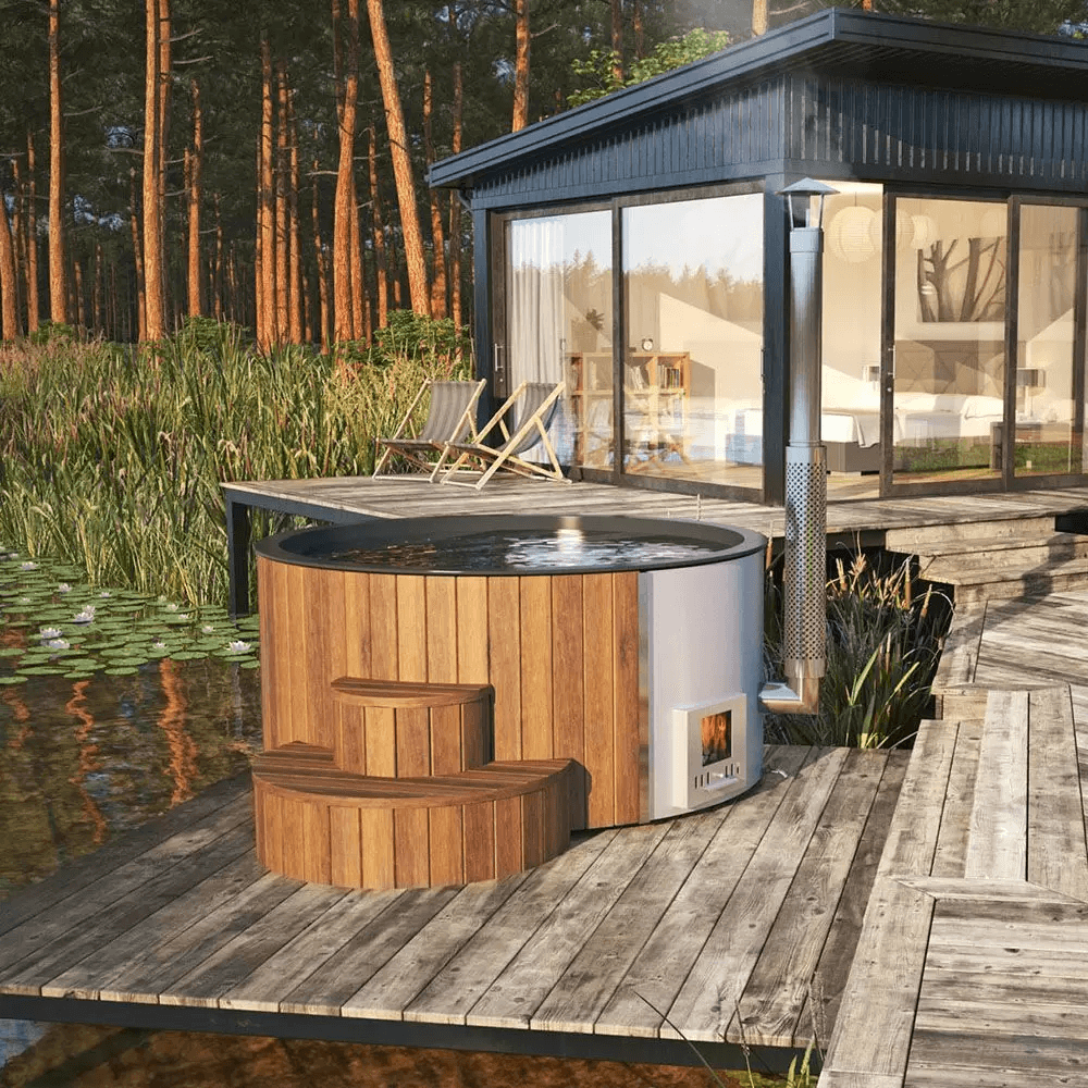 InMedias Delux 220 XL Hot Tub on a rustic wooden deck beside a modern cabin near a lake, surrounded by reeds and forest in daylight.