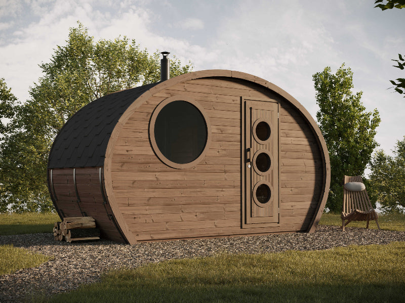 Outdoor photo of the InMedias Frodo Camping 195 pod with a round window and chimney, placed on gravel and surrounded by trees under a cloudy sky.