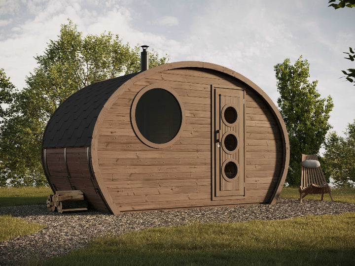 Outdoor photo of the InMedias Frodo Camping 195 pod with a round window and chimney, placed on gravel and surrounded by trees under a cloudy sky.