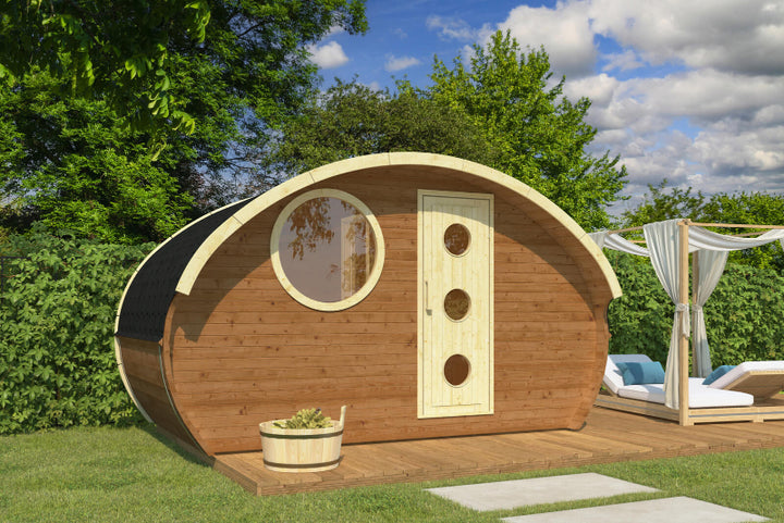 Front view of the InMedias Oval Sauna Frodo 240 with a cream-colored door and circular window, on a sunny lawn with white curtains and lounge chairs , outdoor background with green trees and blue sky.