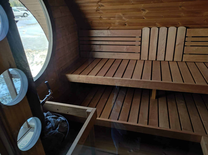 Interior shot of InMedias Oval Sauna Frodo 300, showing wooden benches and round window  and on a natural daylight background.