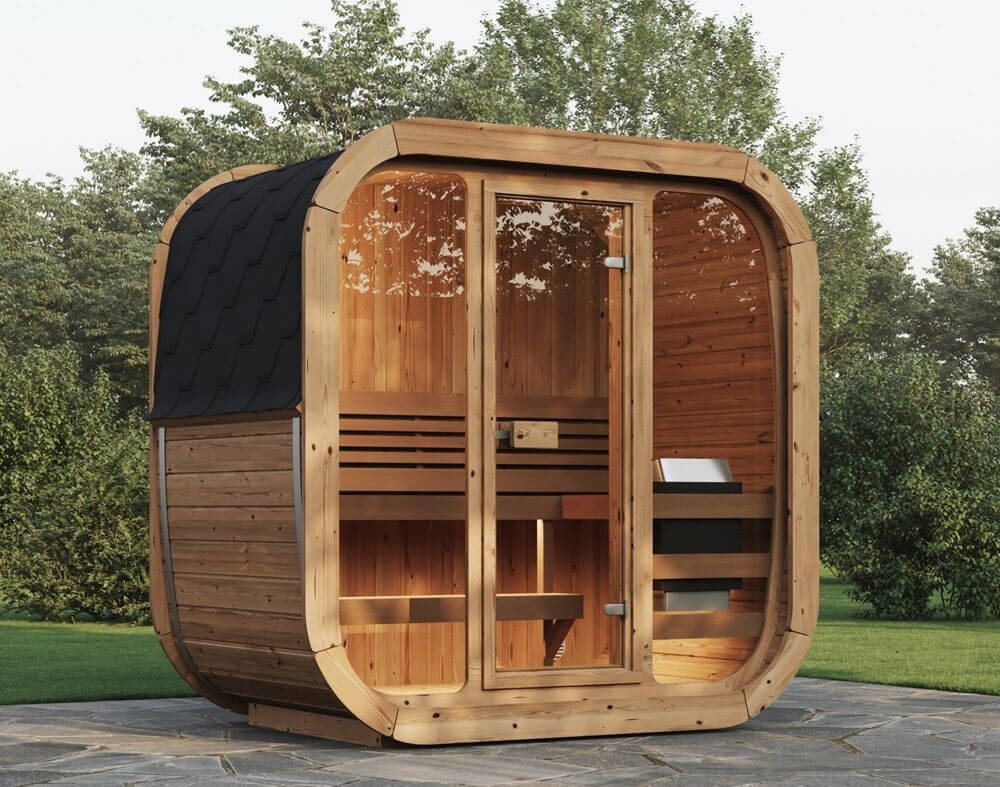 InMedias Sauna Icon 125 full height front view – modern cuboid sauna with glass wall and black shingle roof, placed on a stone patio surrounded by trees, on aoutdoor garden background.