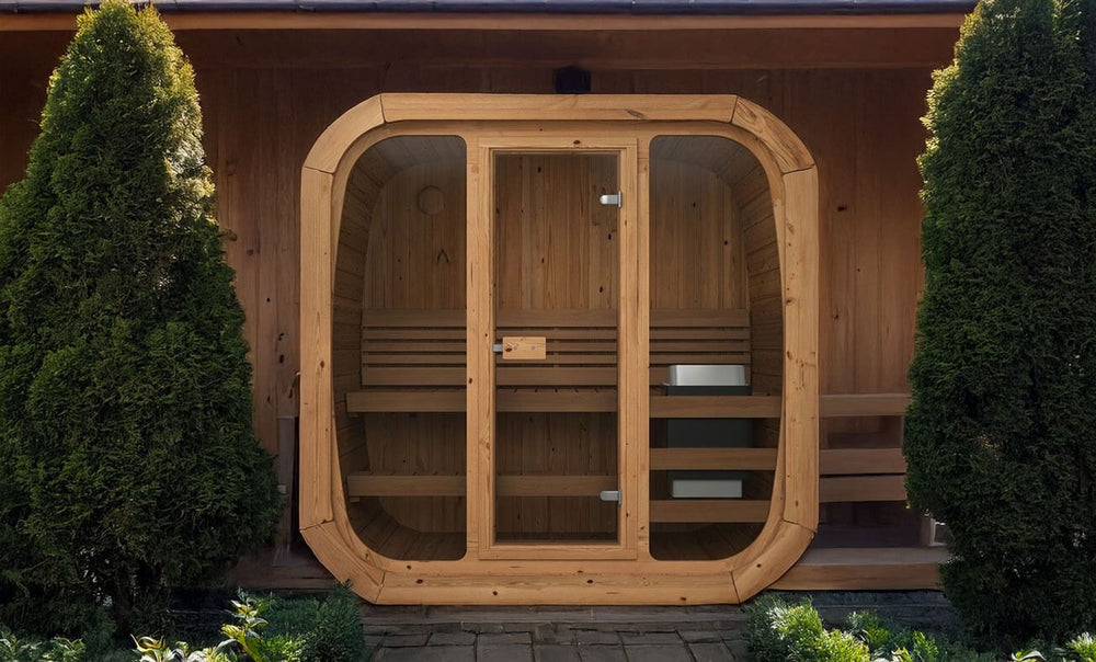 InMedias Sauna Icon 125 front view – a compact wooden outdoor sauna with a full glass front door and side panels, nestled between two trees, on a natural stone path – outdoor background.