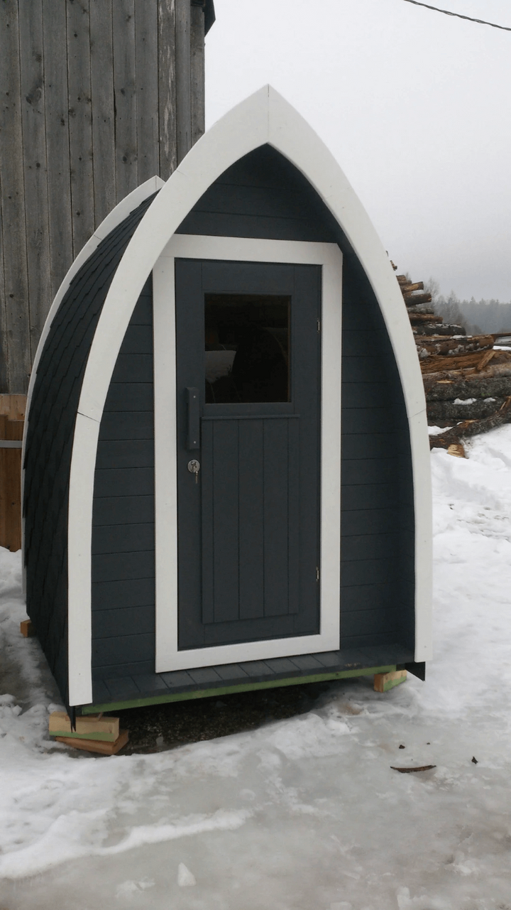 InMedias Toilet Barrel standing on icy ground with dark gray door and white trim, snow-covered setting beside a wooden cabin – wintry outdoor background.