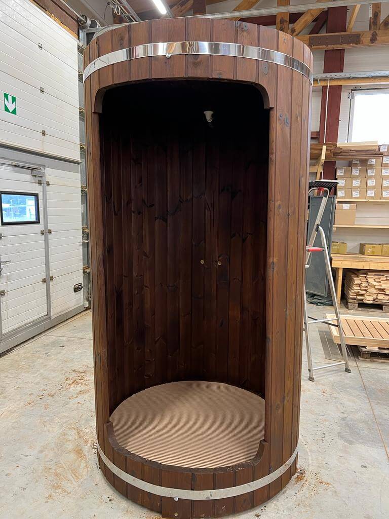 Dark wood InMedias Shower Barrel standing in a warehouse – showing the full structure with chrome bands, on a beige concrete background.