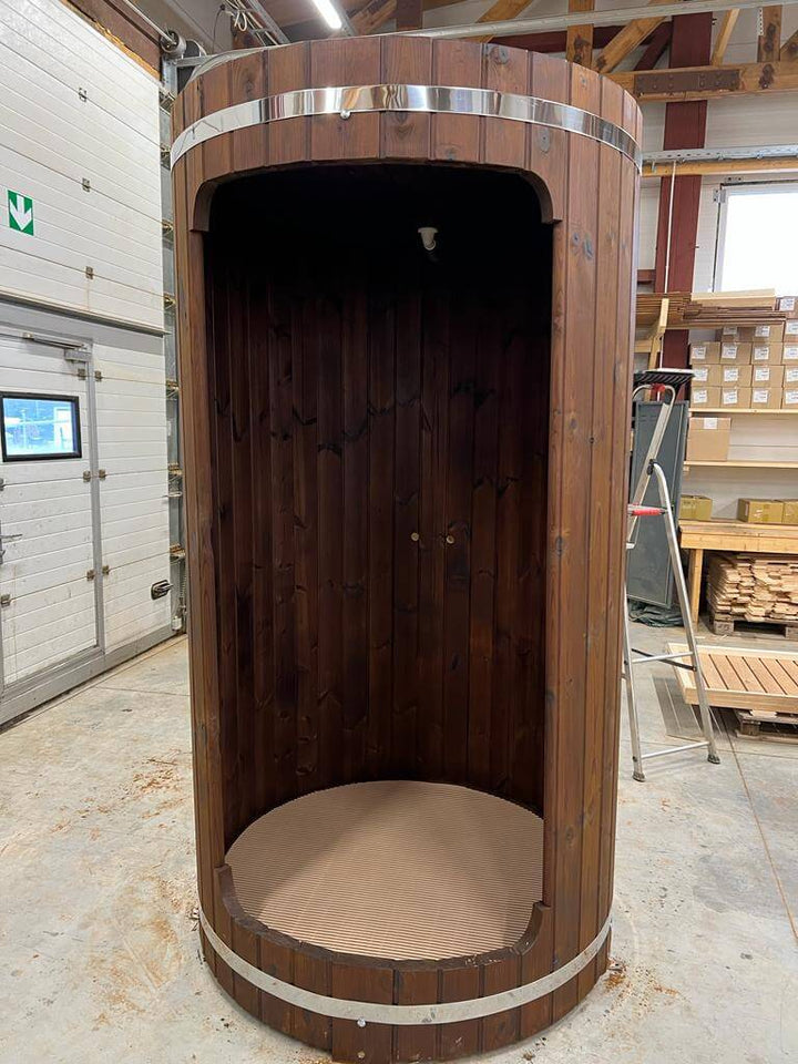 Dark wood InMedias Shower Barrel standing in a warehouse – showing the full structure with chrome bands, on a beige concrete background.