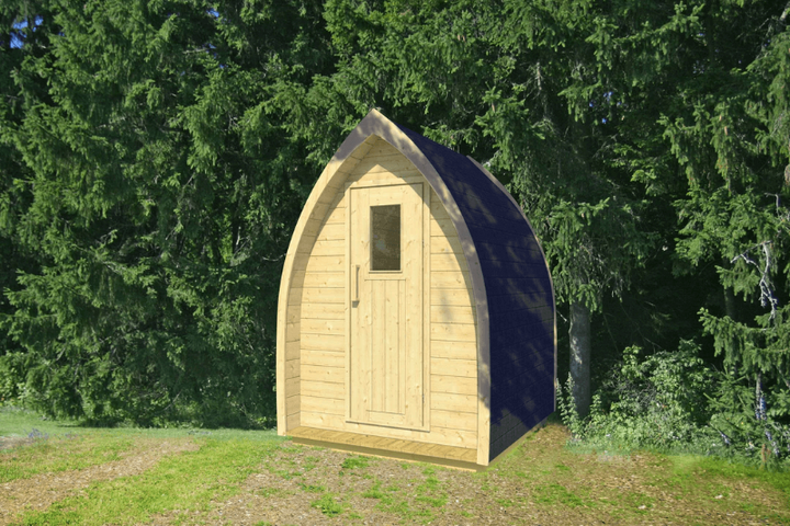 InMedias Toilet Barrel small wooden cabin with a pointed roof and shingled black top, standing on grassy terrain with a forested background – natural daylight, green background.