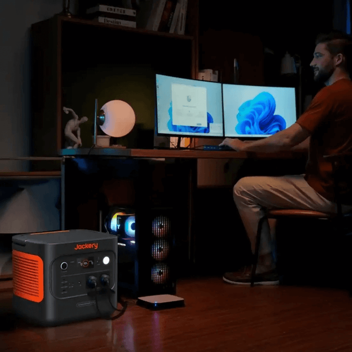 Jackery Explorer 2000 V2 portable power station powering dual monitors in a home office setup with wooden flooring and shelves.