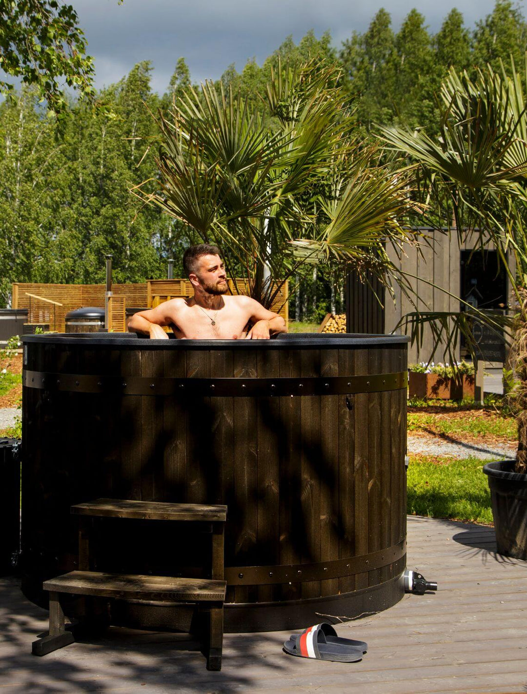 Man relaxing in a Harvia Legend black wooden hot tub outdoors surrounded by trees and garden decor.