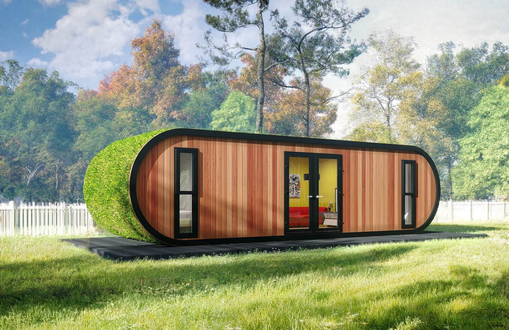 Glamping pod with rounded green-covered end, wooden cladding, and black-framed windows, placed on a platform in a garden with trees and a white fence.