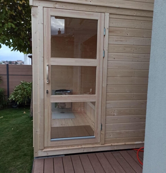 Viking Industrier Sauna Cube 2 x 2m with full glass door on a wooden deck, placed beside a modern house, with green grass and trees in the background.