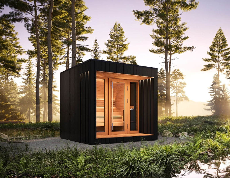Saunum AirCube Terrace sauna placed in a forest setting at sunset, featuring black vertical wood exterior and illuminated wood-lined interior.