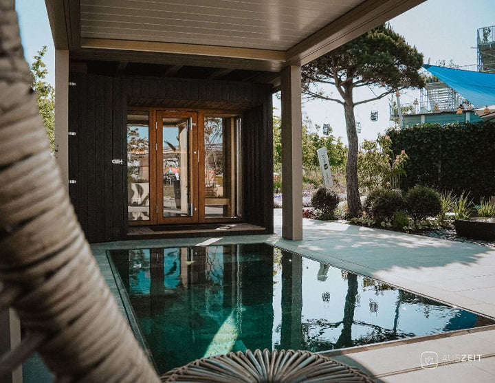 Saunum AirCube Terrace sauna installed beside a private pool under a covered patio, with glass doors reflecting the garden surroundings.