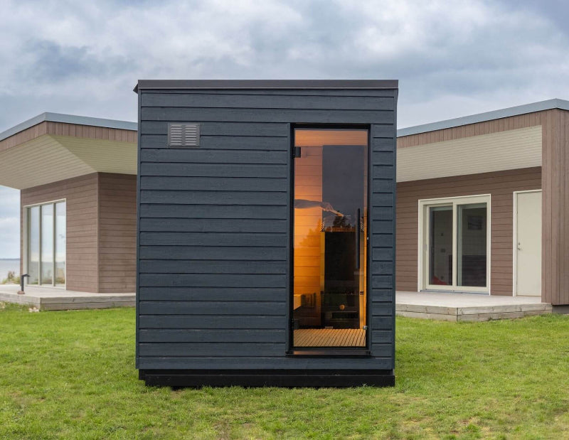 Side view of the sauna showing a narrow vertical window with a glimpse of the heater inside, set against a modern home backdrop.
