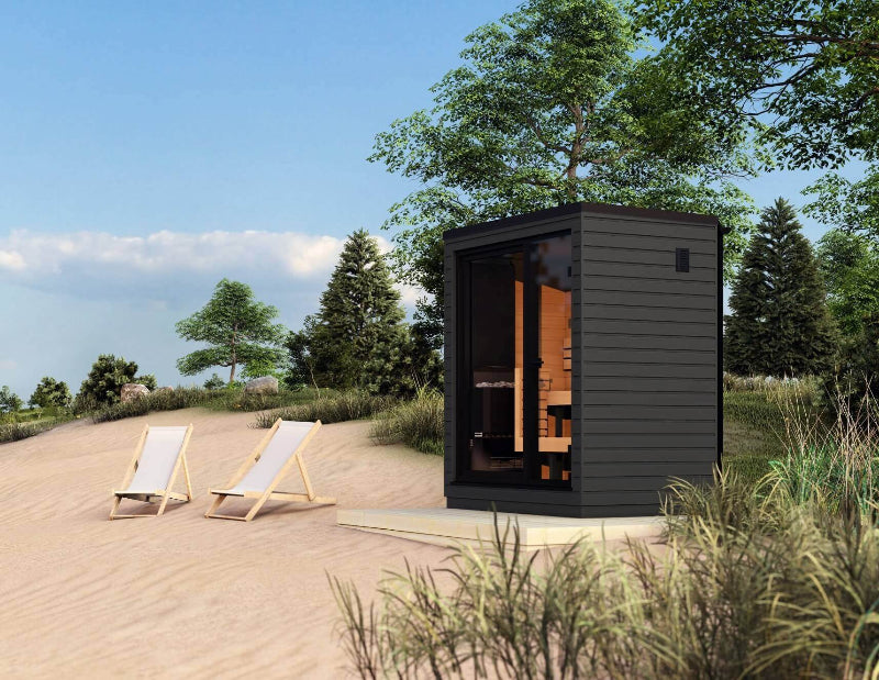 A compact black outdoor sauna cabin on a sandy area with two lounge chairs nearby, surrounded by trees and greenery.