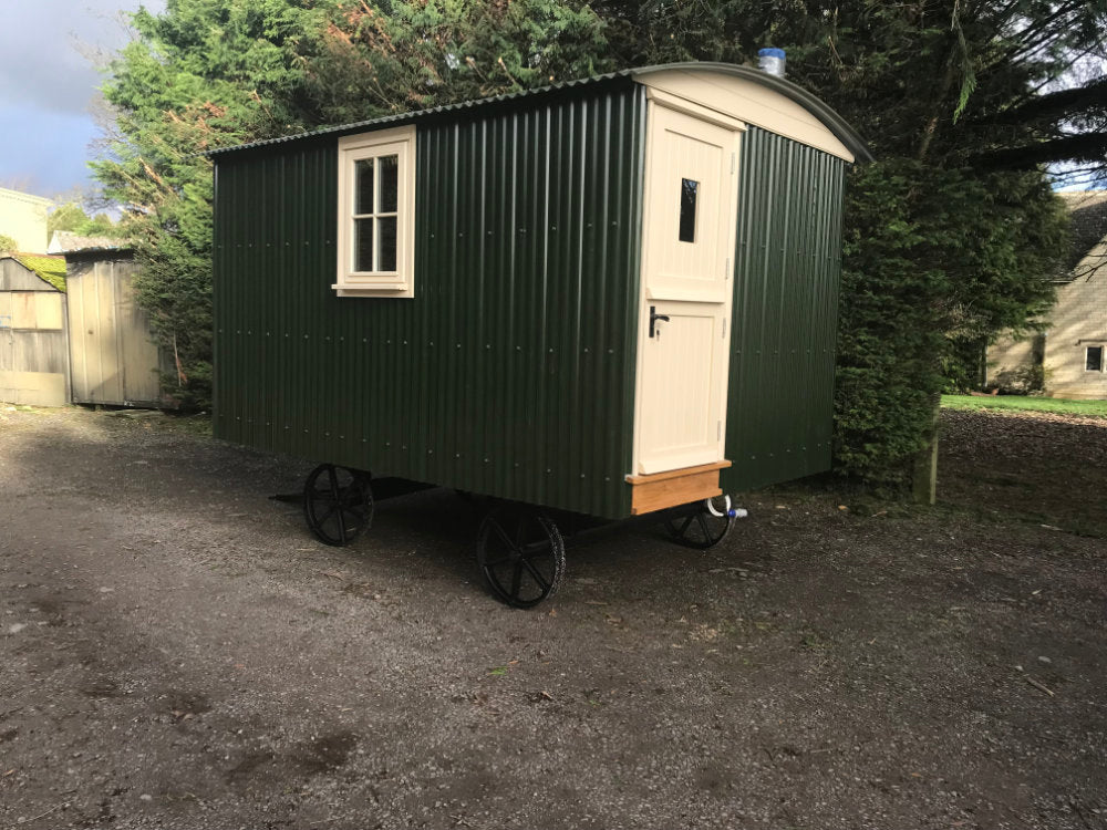 Green corrugated steel shepherd's hut with white door and window, mounted on black cast-iron wheels, positioned on gravel with trees in the background.