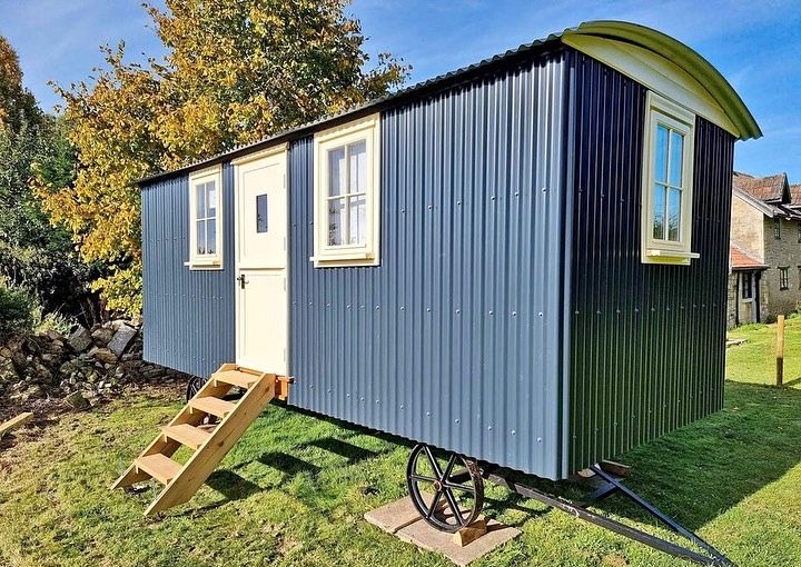 Dark blue corrugated metal shepherd's hut with cream windows and door, placed on a grassy lawn with wooden steps and iron wheels, under a clear blue sky.