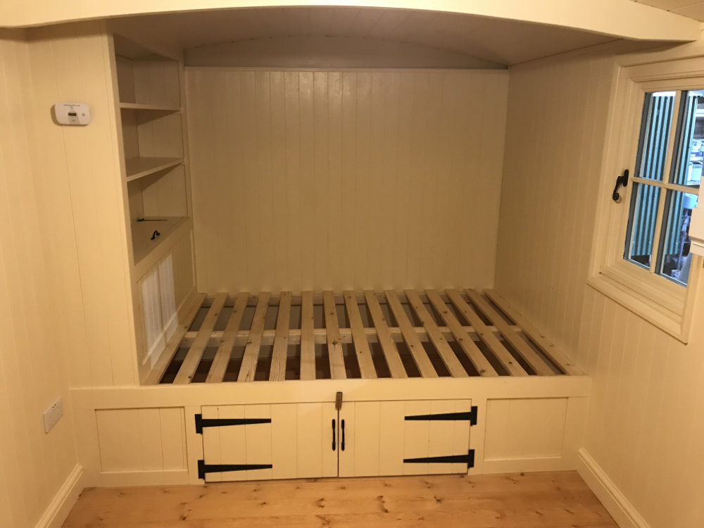Interior of a cream-painted shepherd's hut featuring a built-in wooden bed frame with under-bed storage cupboards, shelving, and a side window.