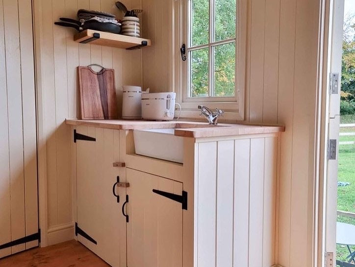 Compact kitchenette inside a shepherd's hut with cream cabinetry, a ceramic sink, wooden countertop, open shelf with dishes, and a window overlooking greenery.