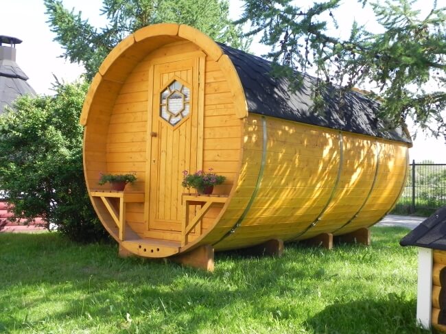 Exterior view of a Viking Industrier 2.2 x 4m Sleeping Barrel with dark shingles and natural wood color, featuring a front door with hexagonal window design, on a grassy outdoor.