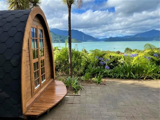 Viking Industrier 2.4 x 3.5m camping pod with rounded wood deck, placed in front of a lush tropical lake view under partly cloudy skies.