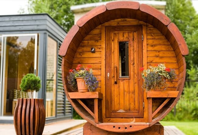 Centered view of the Viking Industrier Barrel Sauna 1.9 x 2.5m with curved wood facade, built-in benches and flower pots, situated on a wooden deck in a garden setting.