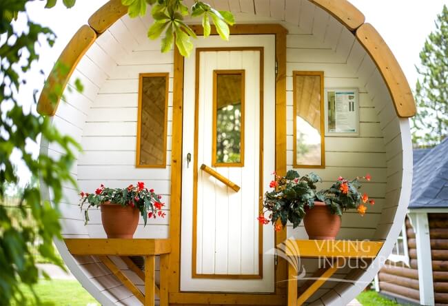 Close-up front view of the Viking Industrier Barrel Sauna 1.9 x 3m in white finish with wooden trim and flower pots on each side, placed outdoors among greenery.