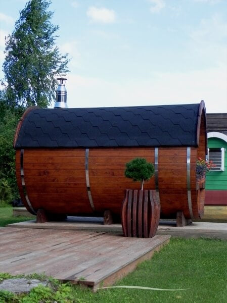 Side view of Viking Industrier Barrel Sauna 2.2 x 2.5m in dark brown finish with black roof shingles, placed outdoors on a wooden deck with a planter.