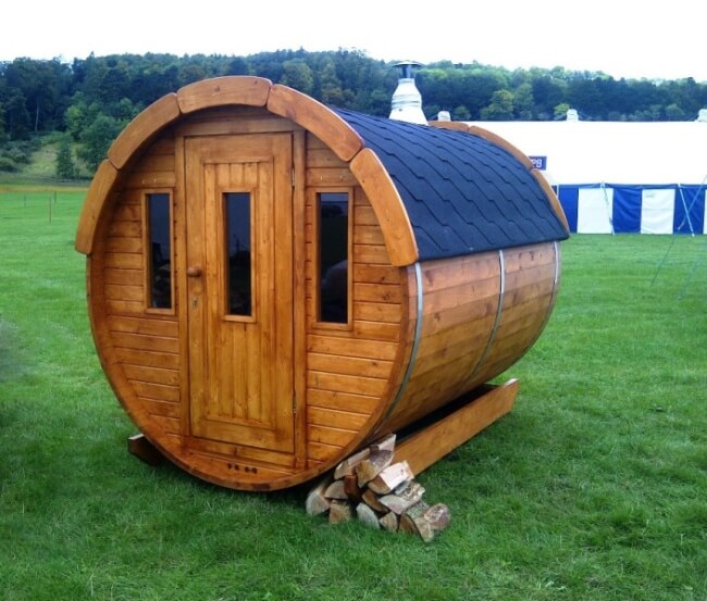 Viking Industrier Barrel Sauna 2.2 x 2m made of natural wood with a black shingled roof and firewood stacked at the base, placed on a grassy outdoor area.