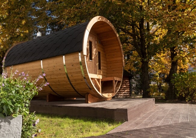 Side view of the Viking Industrier Barrel Sauna placed on a raised platform in a green garden, with autumn trees in the background.