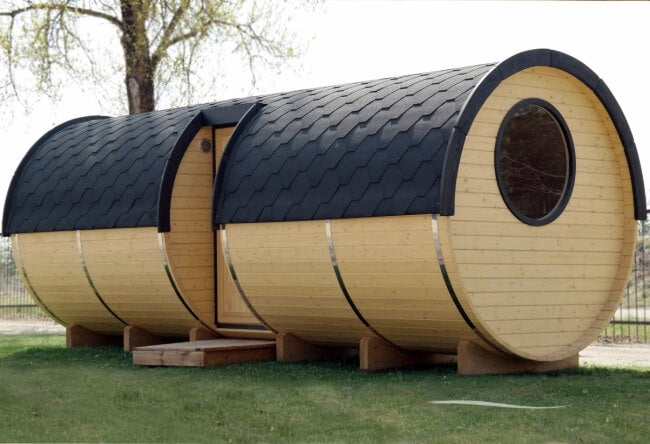 Exterior side view of Viking Industrier Barrel Sauna 2.2 x 5.9m with a round window, dark shingle roof, and natural wood finish, situated on green grass.