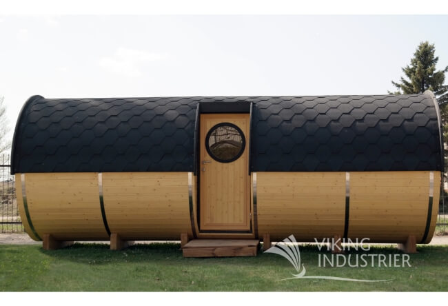 Front view of Viking Industrier Barrel Sauna 2.2 x 5.9m with side entrance, showing circular window on wooden door and black shingle roofing, placed on a grassy lawn.