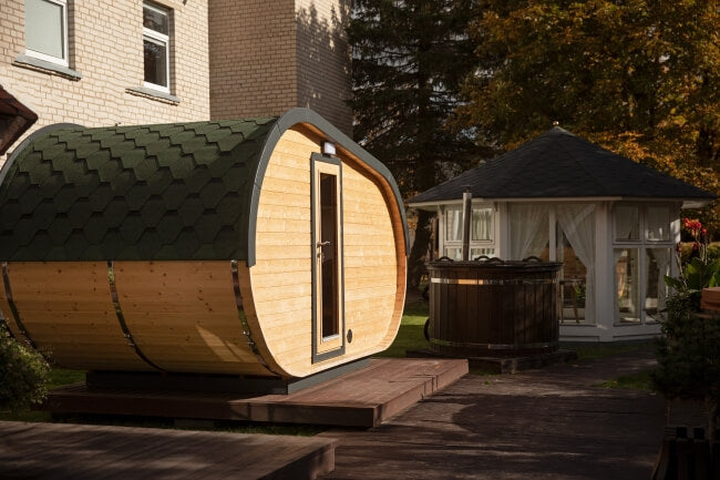 Front view of the Viking Industrier Delight Camping Pod 2.4 x 4.3m with green roof and natural wood walls, positioned in a garden next to a white gazebo and hot tub. Background includes house and trees.