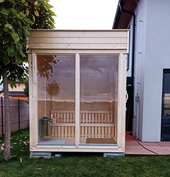 Viking Industrier Sauna Cube 2 x 2m viewed from the front, showing a large full glass window and wooden bench interior, located outdoors with a tree beside it.