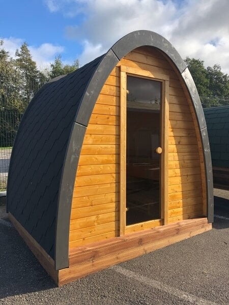 Angled front view of Viking Industrier Sauna Pod 2.4 x 2.3m with black shingle roof and glass door, on a paved outdoor surface.